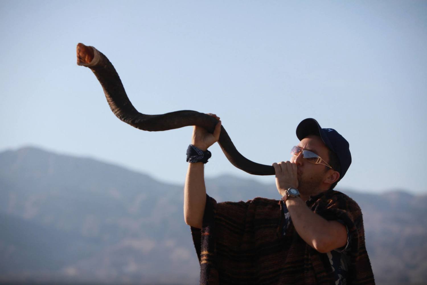 A Jewish man in sunglasses and a baseball cap and a woven tunic with magical wrist charms on playing a large ram's horn shofar in the desert in front of a mountain range