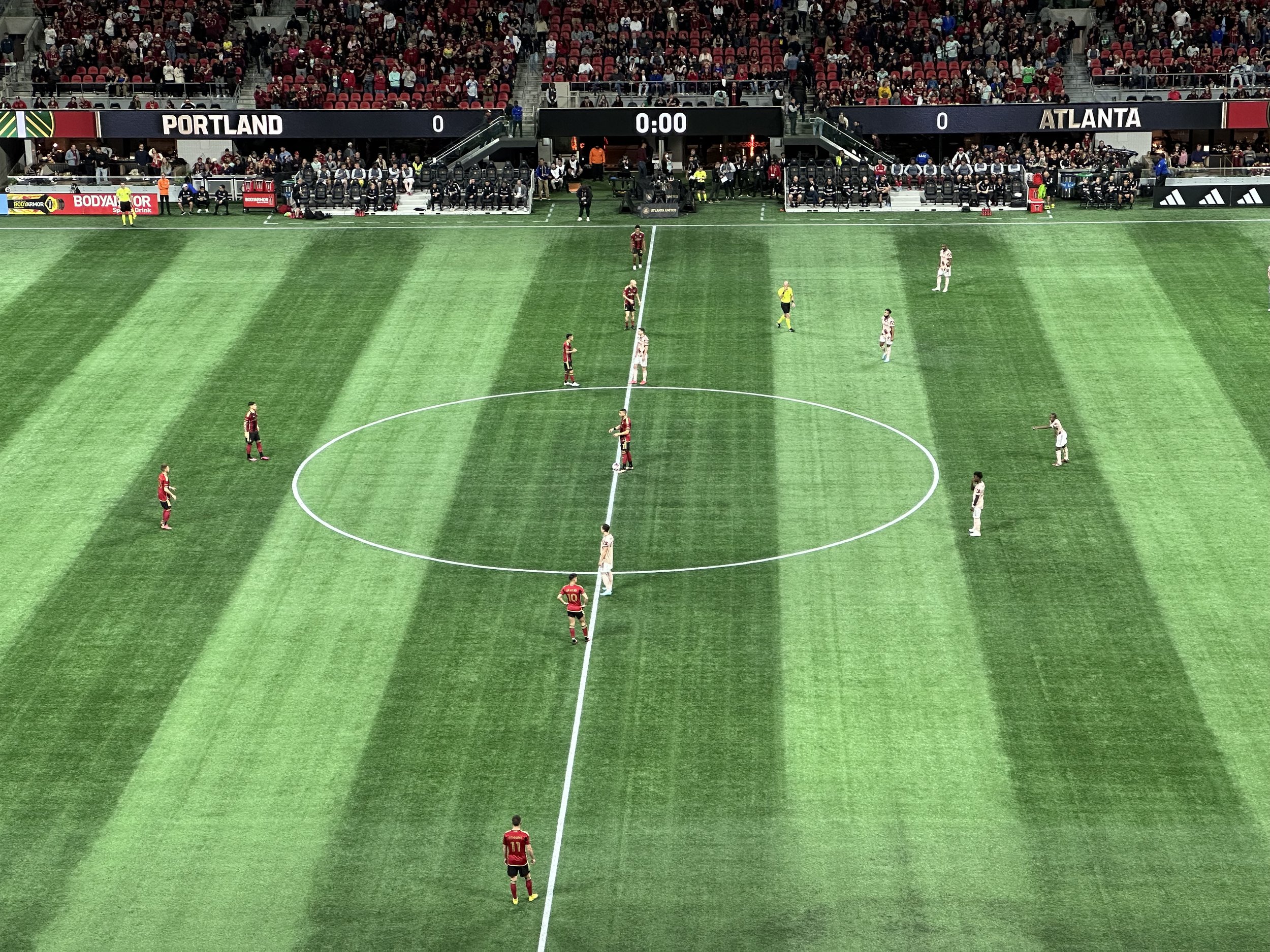 A photo of midfield at a soccer game just before kickoff, with players on both sides assembling. The scoreboard on the other side of the field reads "Portland: 0, Atlanta 0," and the play clock reads "0:00."