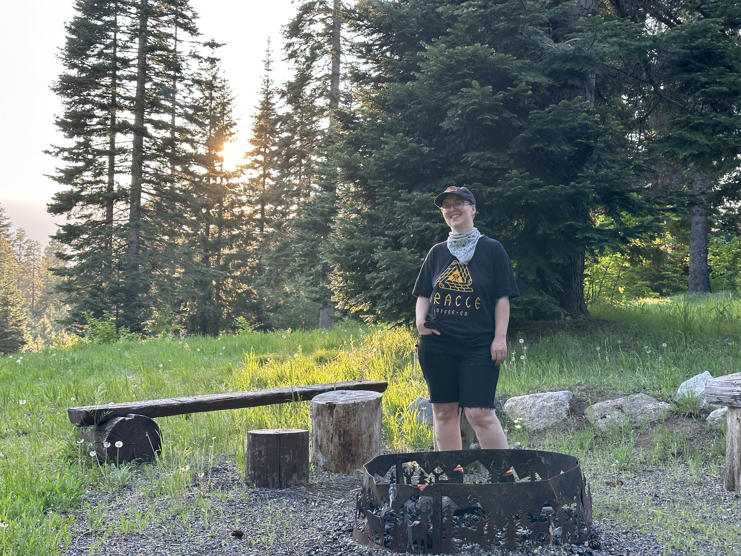 A person in a black cap, shorts, and t-shirt that says "ORACLE," standing in a fire pit in front of evergreen trees and the setting Sun.