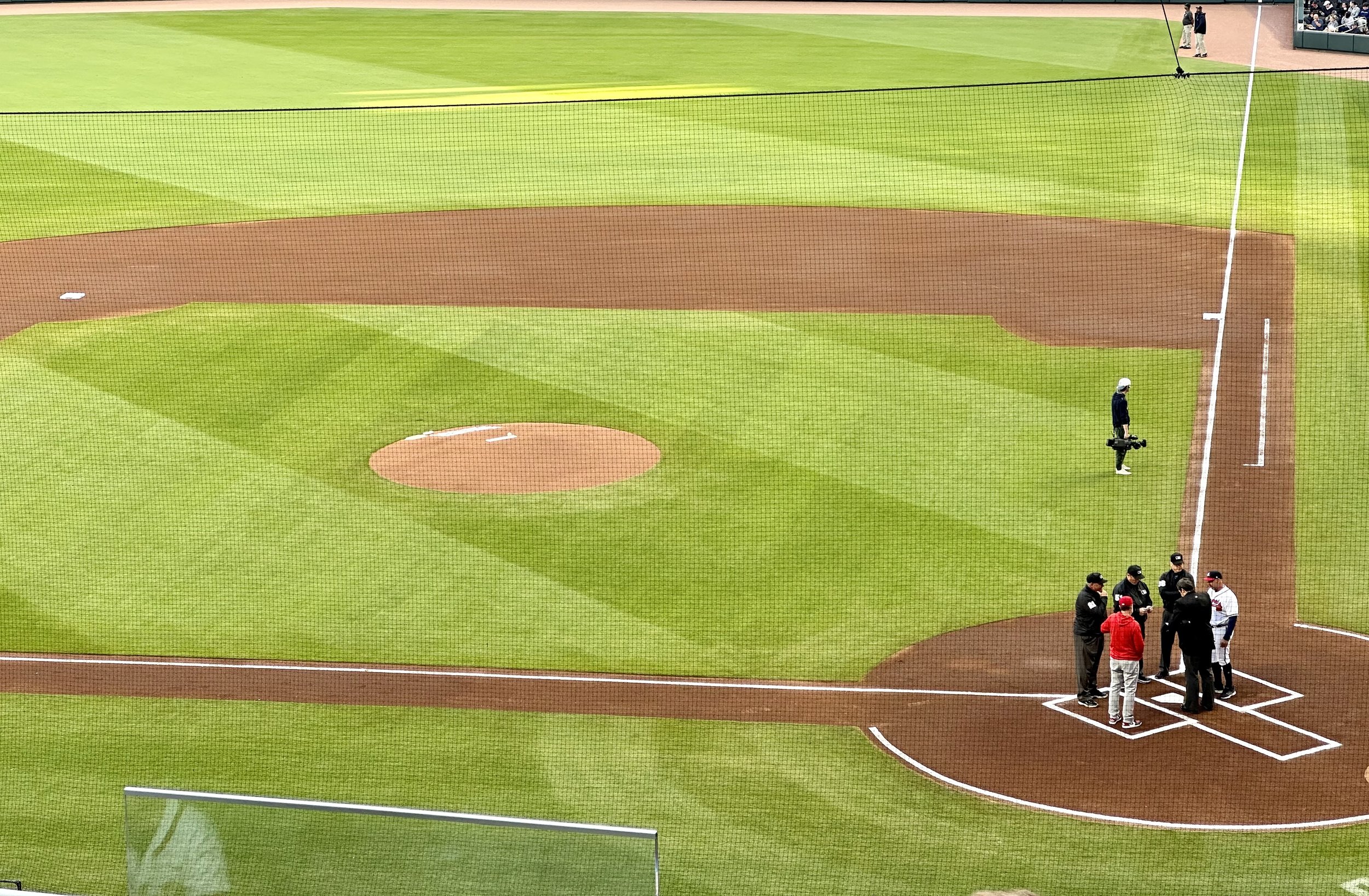 A baseball diamond viewed from seats on the 3rd base side of home plate, with umpires and managers gathered around the plate before the start of the game.