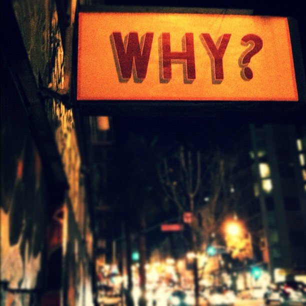 An urban street scene with a large orange rectangular sign overhead that says "WHY?" in large red letters