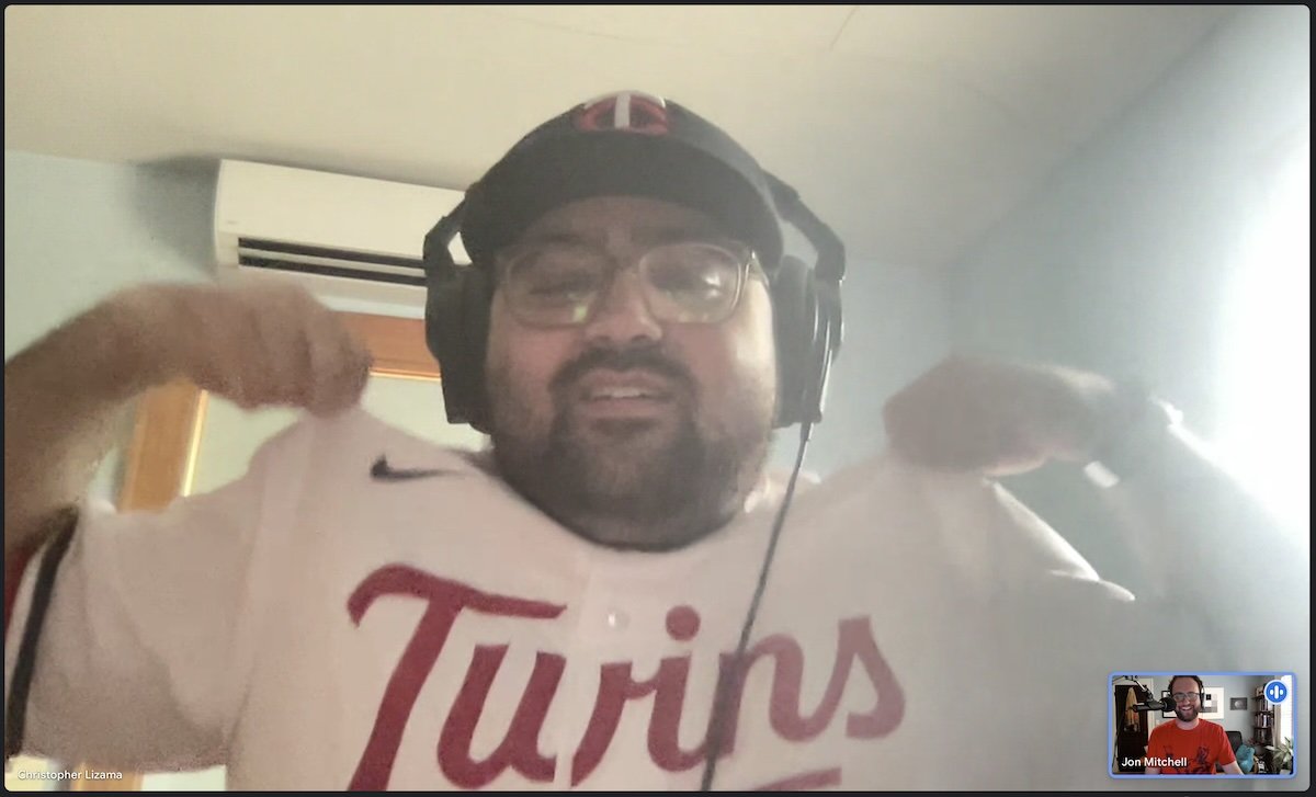 Two people talking on a video call. The main person is wearing full Minnesota Twins regalia. He has on over-ear headphones over his cap. He is showing his jersey to the camera. The other person in the small box is smiling.
