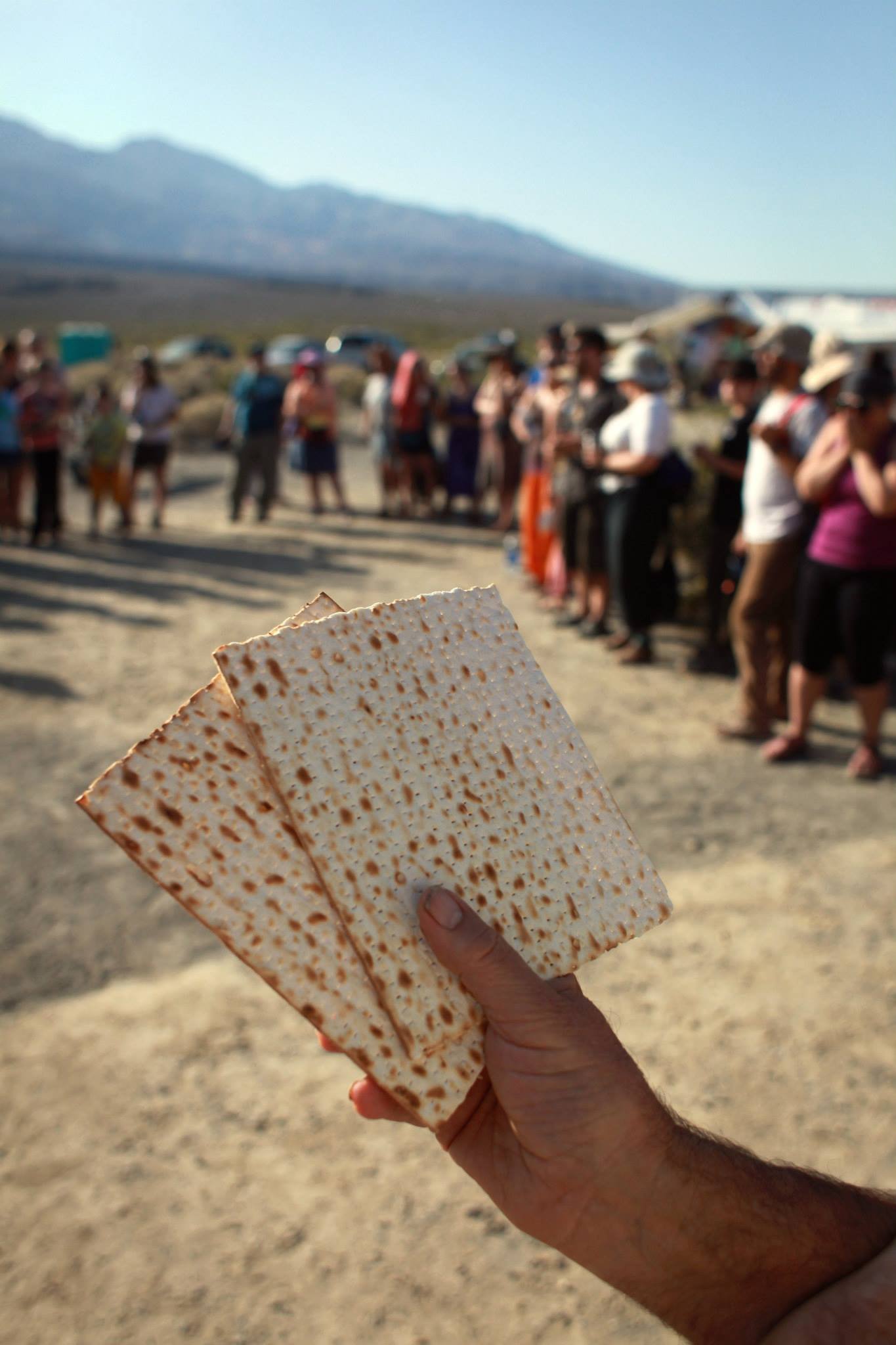 A hand holding two square pieces of matzah at a pre-meal circle gathering of Jewish people in the desert.