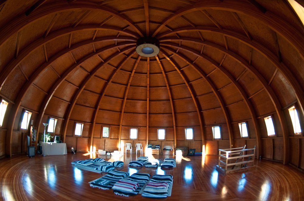 A domed wooden chamber that looks like the interior of a ship, with arched beams leading to a black circular aperture at the apex. Rectangular portholes surround the room at eye level. There are striped woolen blankets on the floor.