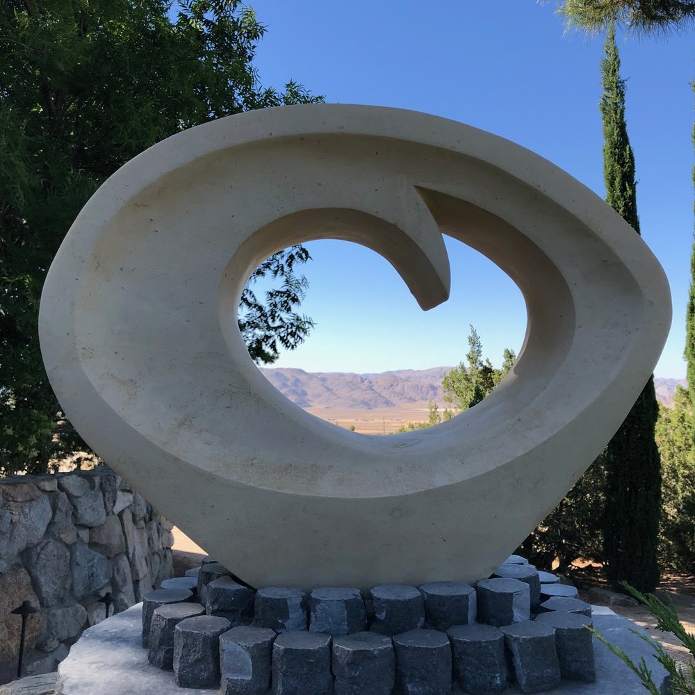An oval stone sculpture shaped like an egg or eye with a hole in the center that has a wave curling into it at the top. It's placed in the garden in a retreat center, and you can see desert and mountains beyond through the hole