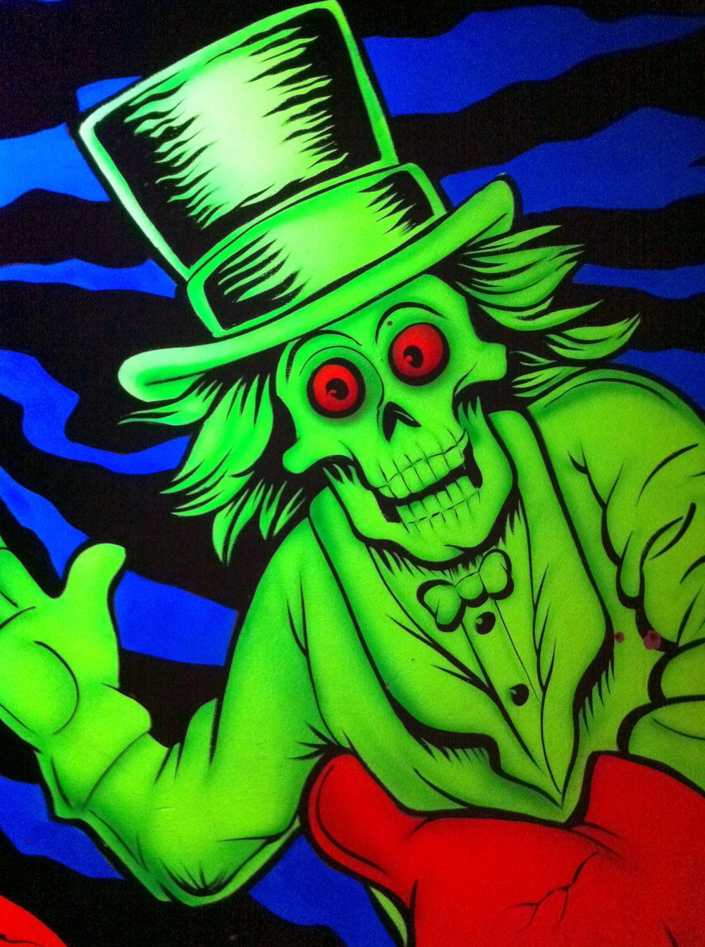 A neon green skeleton with red eyes and a full head of hair dressed in a tuxedo and top hat in front of a field of black and blue stripes