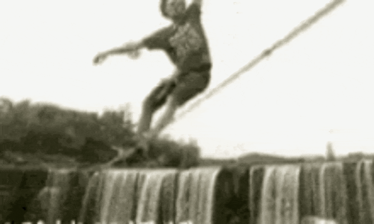 A skinny young white man with frizzy hair in a t-shirt and shorts tightrope-walking across an enormous waterfall wobbling back and forth madly but not falling down.