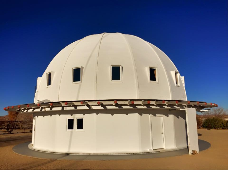 A two-story white dome with vertical rectangular windows and a ring of wooden beams with red tips sticking out horizontally at regular intervals. There is a narrow concrete sidewalk around the structure and a closed door with a doorknob.