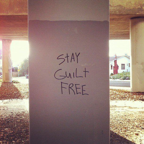 A concrete support column under a highway overpass with the words "Stay Guilt FREE" written on it in permanent marker