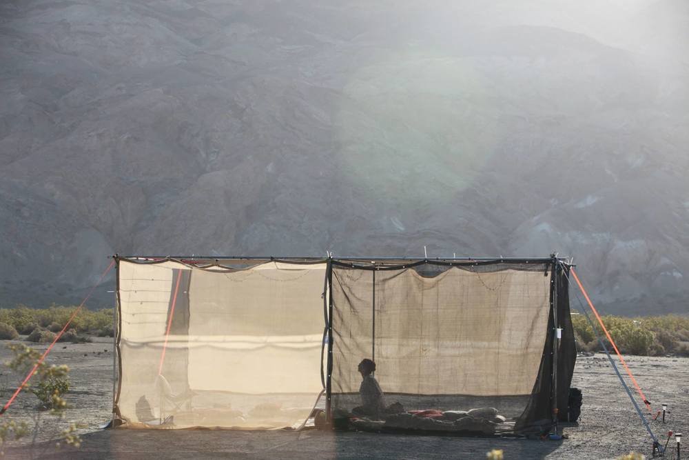 A temporary desert structure with a nearby mountain range in the background with the rising sun streaming over the top. One person is praying inside the structure, which has fabric walls that can be seen through.