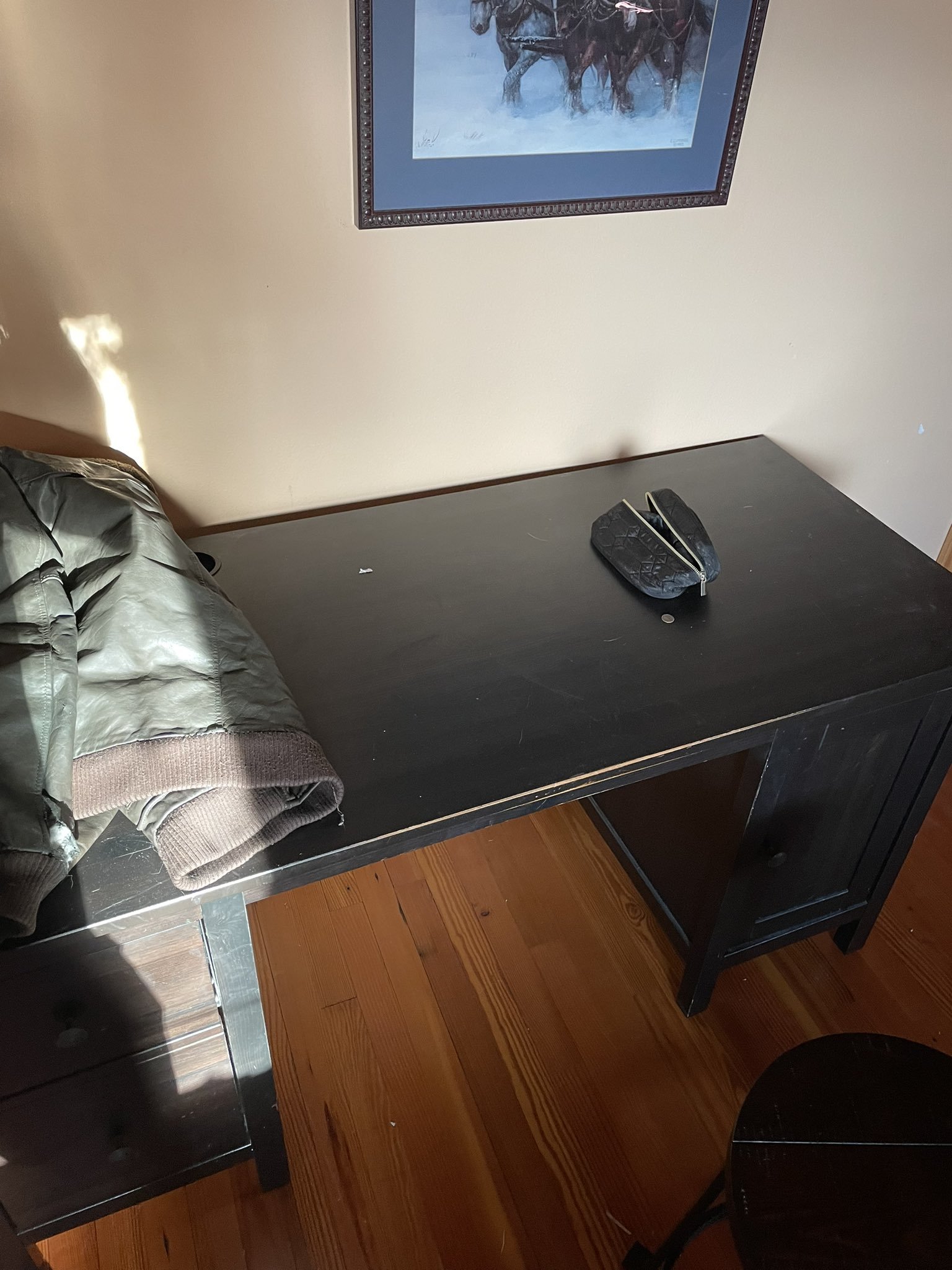 Photo of a black desk against a beige wall on a wood floor. A framed picture of horses in the snow hangs above the desk. On the desk there is a folded black leather jacket and a leather zippered pouch with a dime lying next to it.