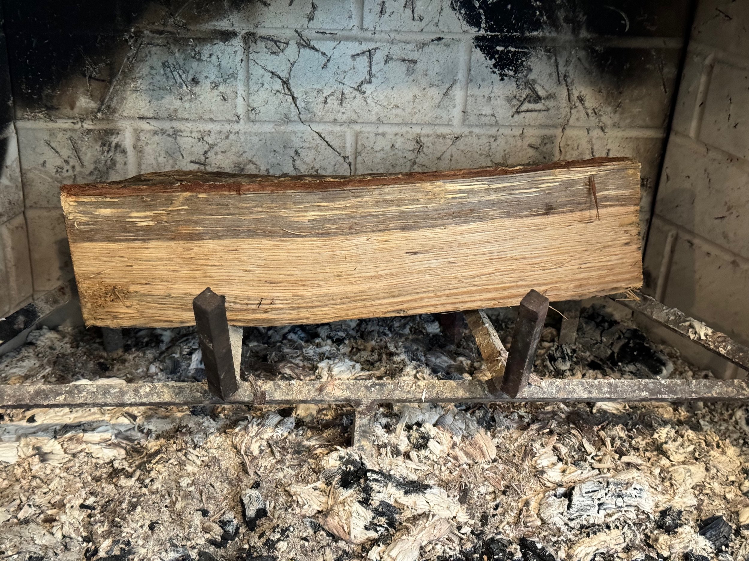 A small fireplace with a metal rack with one log with a cleanly cut straight side facing front from the back. There is a decent bed of ashes on the floor of the fireplace.