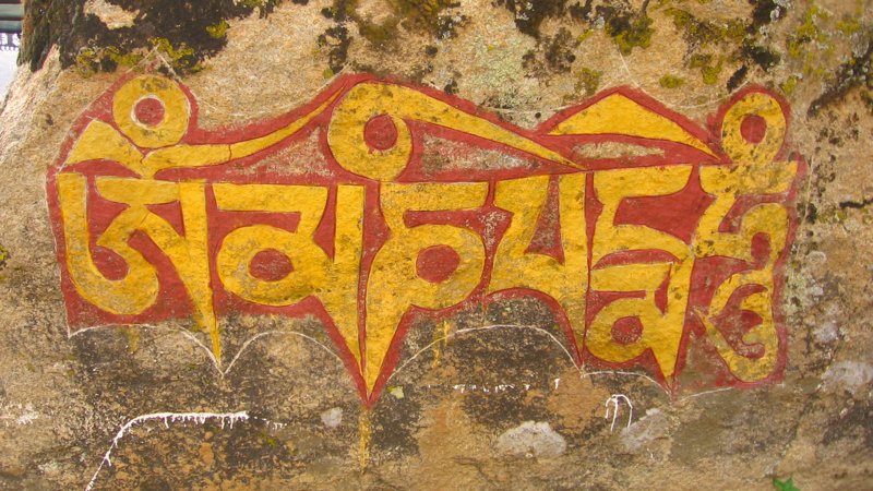 A large lichen-spotted rock with the "OM MA NI PE ME HUNG" mantra painted on it in yellow Tibetan writing with red outlines