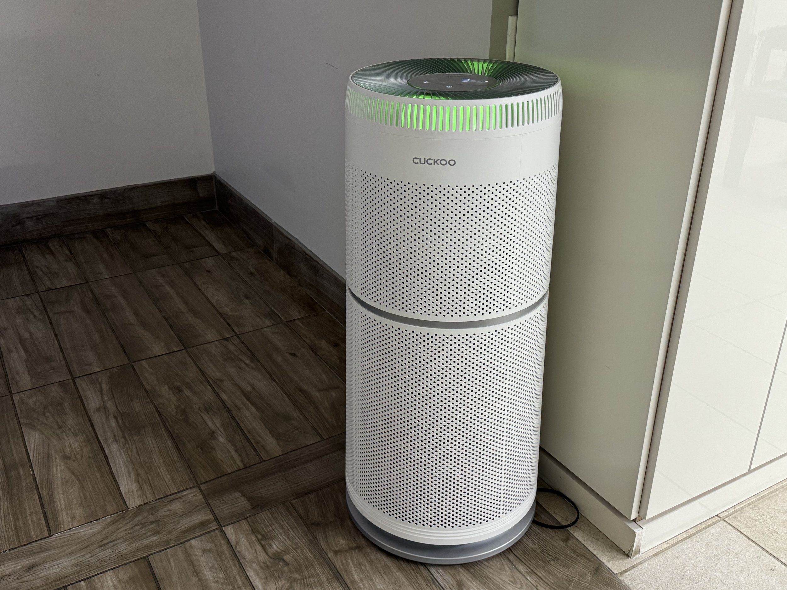 A white air filter robot with eerie green lights inside. The brand is Cuckoo. It's a white cylinder with perforations all over it standing about three feet tall. It's on a fake wood floor in front of a blank white wall.