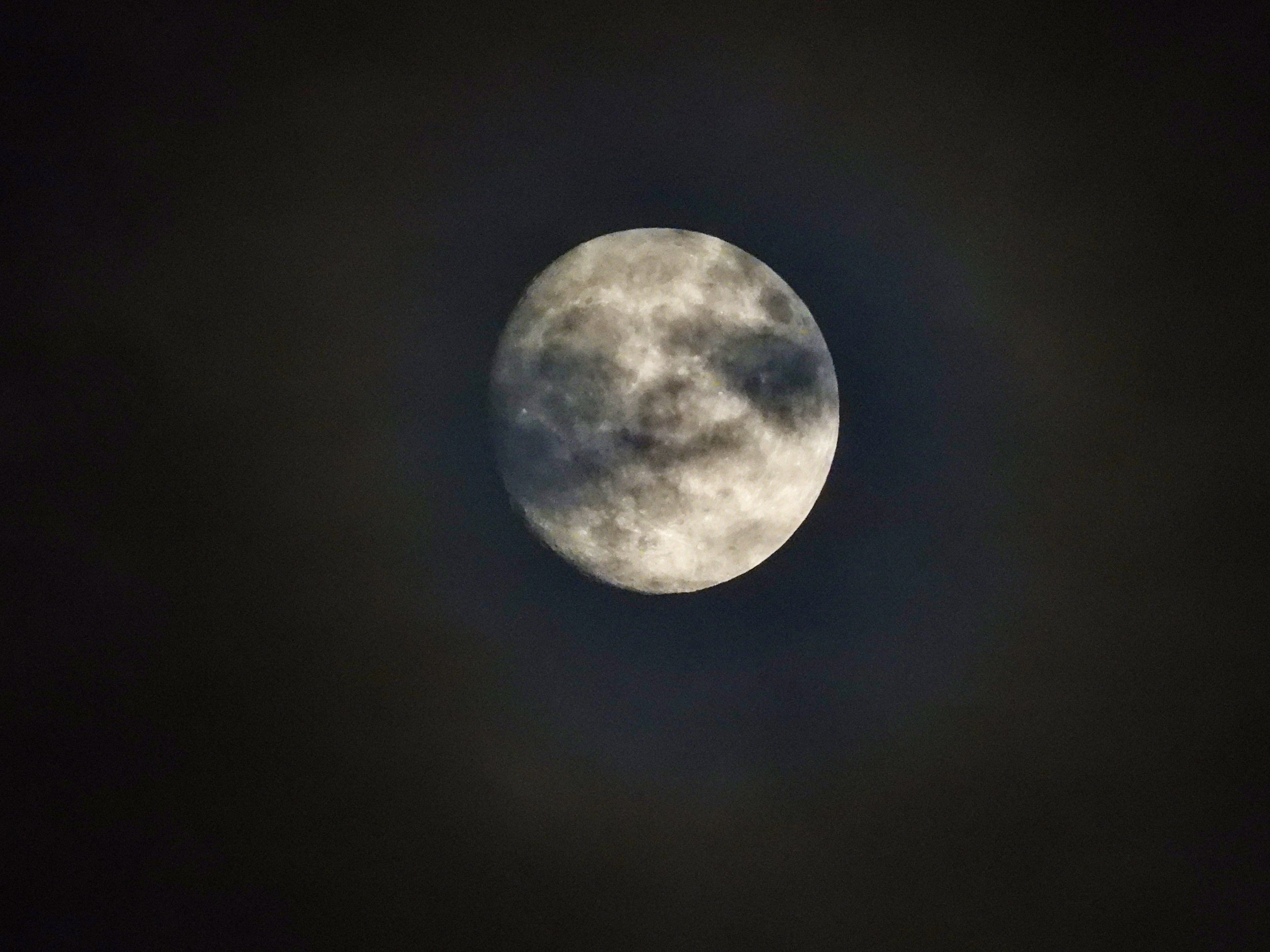 A waxing gibbous Moon pretty blurry and obscured by clouds but with some detailed craters visible on the edges. A dull misty halo is lit up around it in amidst the murky clouds.