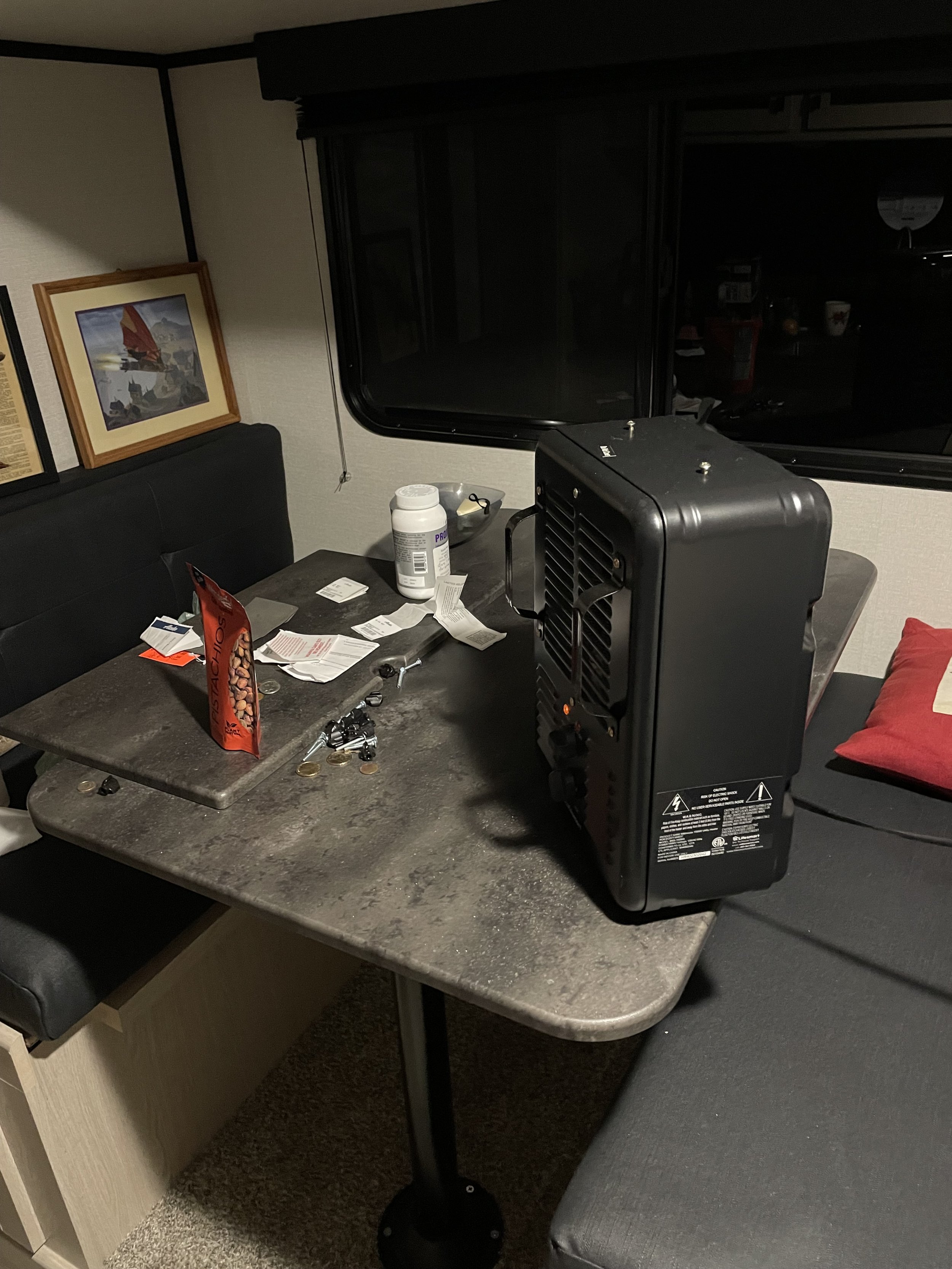 Photo of a dark colored junk-laden table in the expanded section of an RV. There is a black space heater on the table alongside receipts, a bag of spicy pistachios, and other random crap.