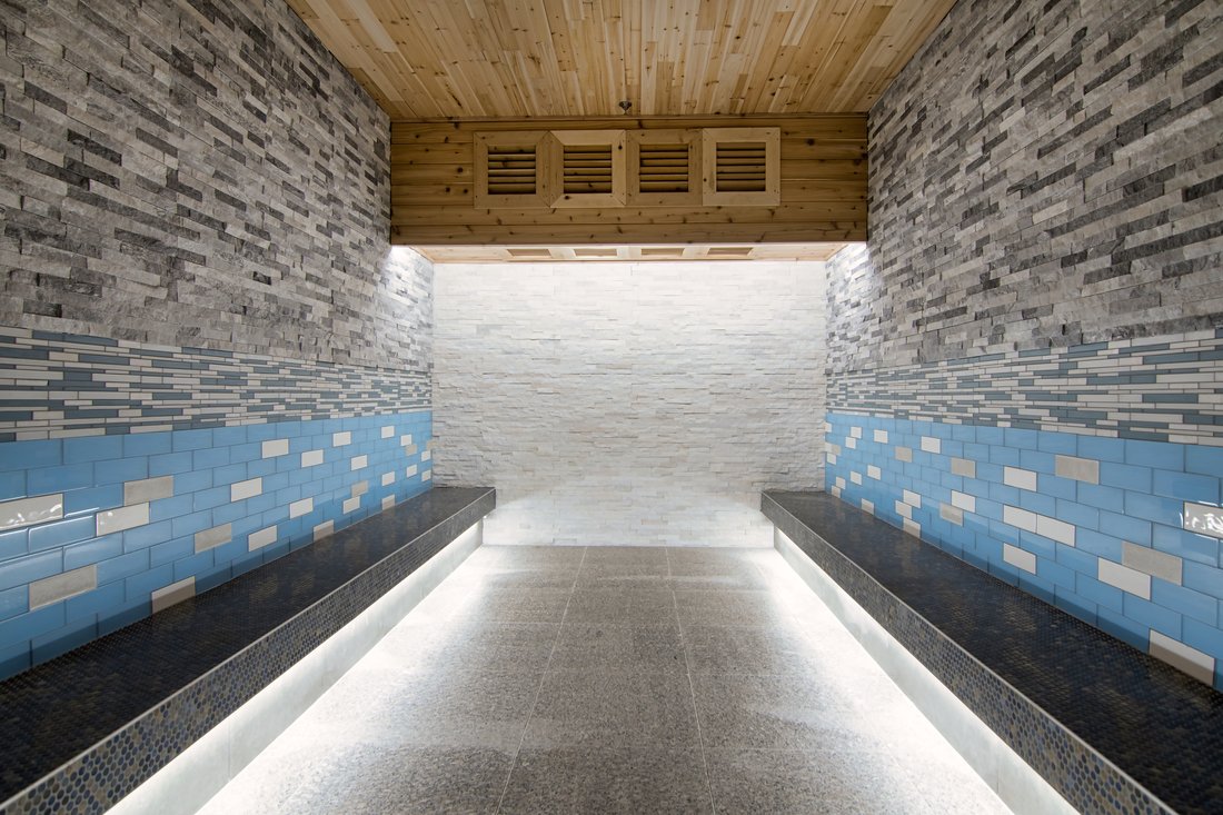 A gray and blue tiled room with large wood-paneled air conditioning ducts and white track lighting on the floor. The sides have tiled benches for sitting.