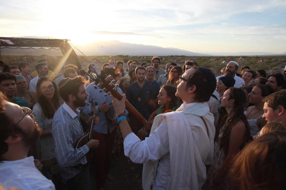 A circle of Jewish people with musical instruments praying in the desert with the Sun setting over a mountain in the distance