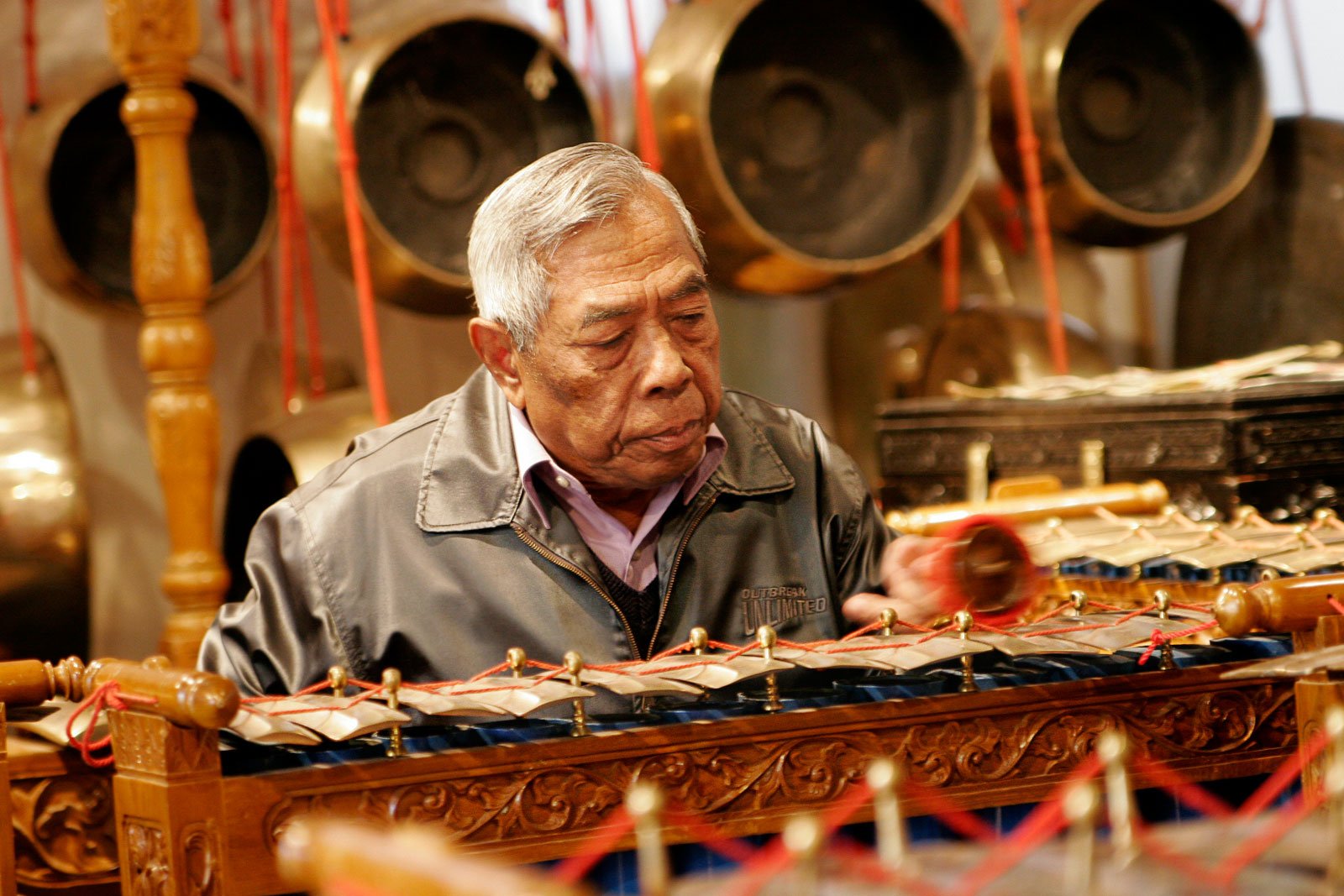 An older Indonesian man in a black leather jacket playing a gamelan instrument surrounded by ornately carved wooden and cast bronze gongs and other instruments.