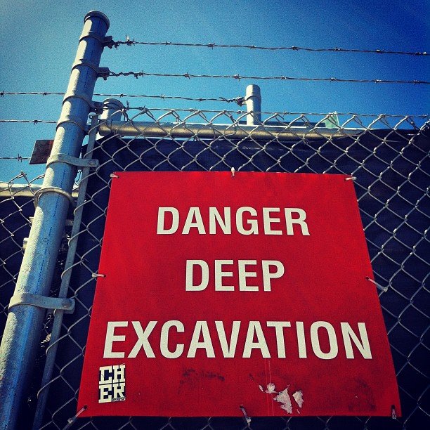 A chain link fence topped with barbed wire with a solid black barrier behind it, with a square red sign on it with white letters that say "DANGER DEEP EXCAVATION"